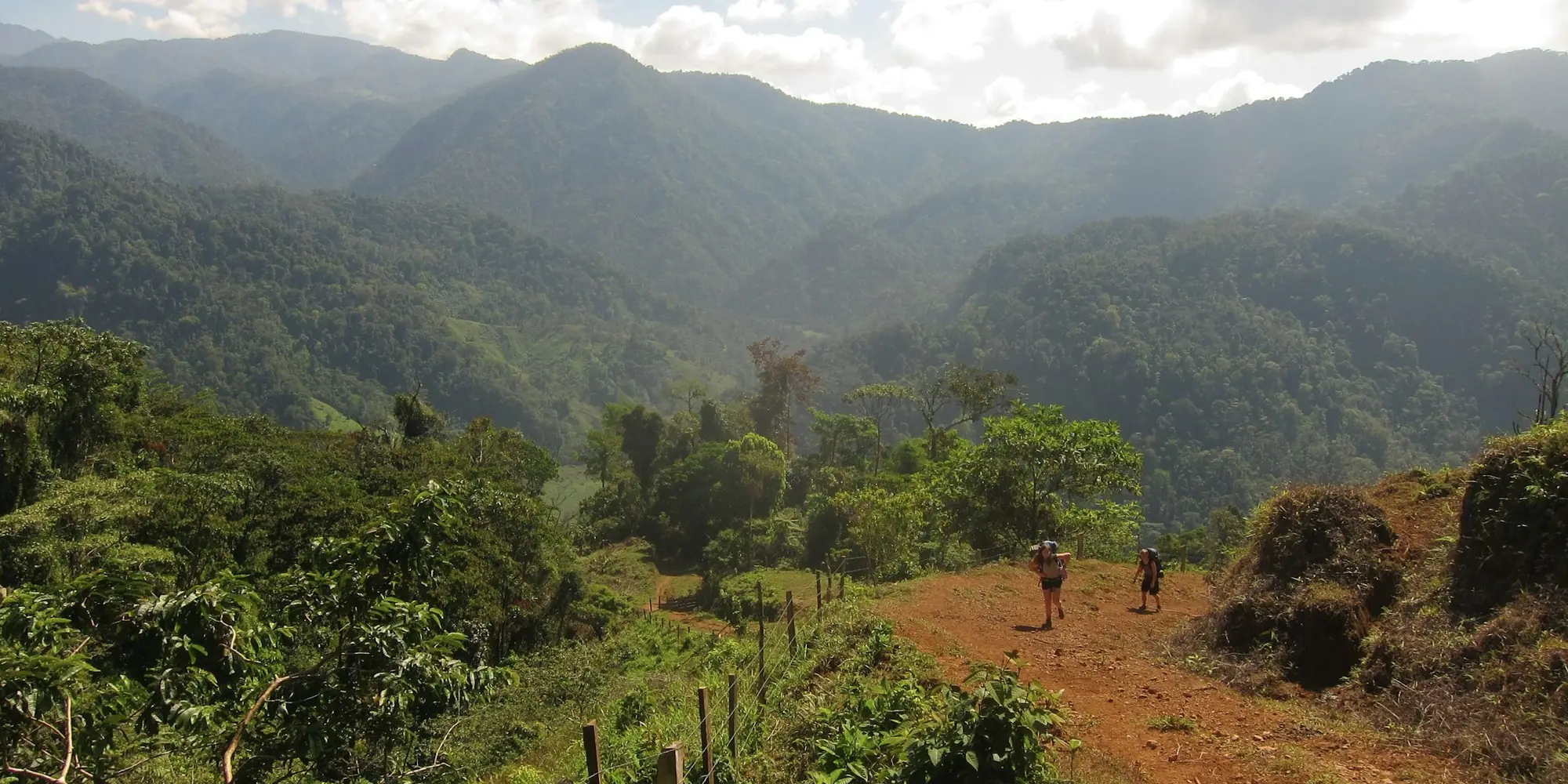Dos personas caminan por un camino de tierra roja junto a una cerca, hacia colinas con bosque en Costa Rica.